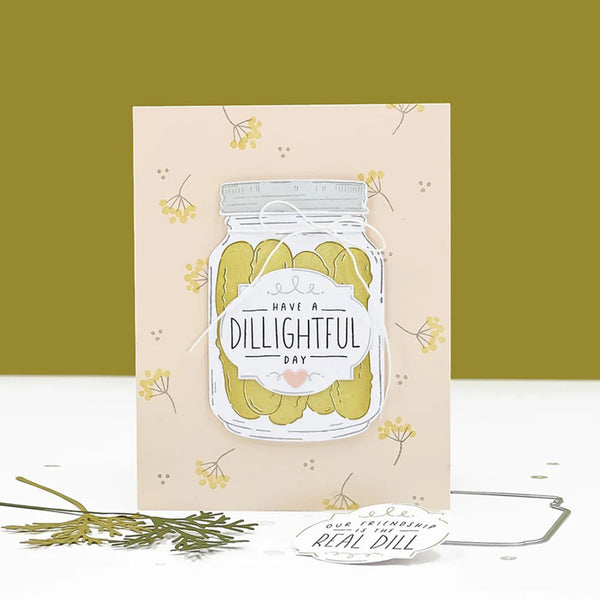 A handmade greeting card featuring an illustration of a glass jar filled with pickled cucumbers and the text 'HAVE A DILLIGHTH DAY'. The card is displayed on a white surface with a yellow-green background, accompanied by a small tag and sprigs of greenery.