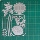 A metal die-cut craft set featuring a mason jar with floral details, a decorative label reading 'HELLO', and a matching label reading 'THANKS', displayed on a green cutting mat with measurement markings.