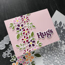 A handmade greeting card with a pink background and layered floral cutouts in pink, purple, red, green, and yellow, alongside silver die-cut metal embellishments on a dark wood surface.