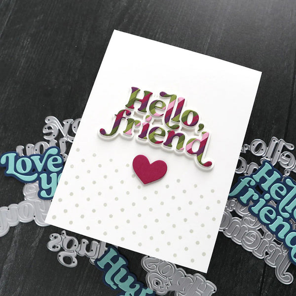A handmade greeting card with 'Hello, friend' in multicolor bubble letters and a small pink heart, displayed on a dark wood background with metal die-cut lettering pieces.
