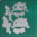 A set of silver metal die-cut craft letters spelling out 'Hello friend', 'Hug', 'You', and 'Hello friend' in multiple sizes, arranged on a green gridded cutting mat.