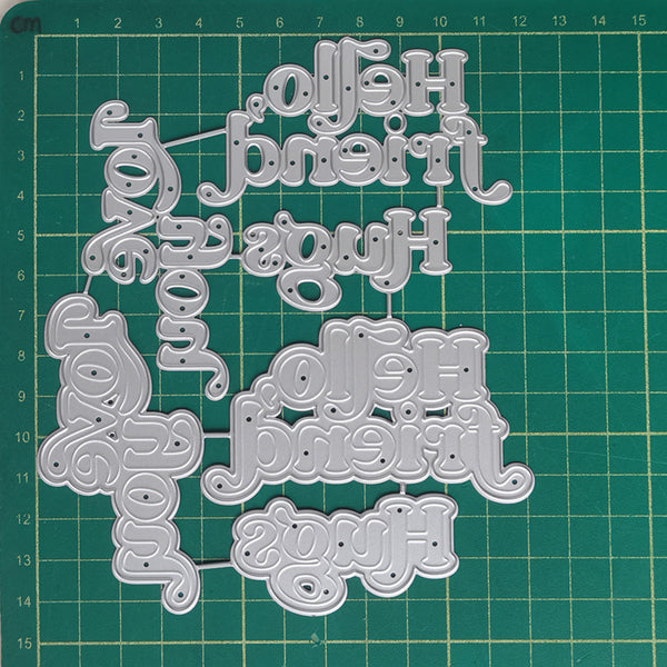 A set of silver metal die-cut craft letters spelling out 'Hello friend', 'Hug', 'You', and 'Hello friend' in multiple sizes, arranged on a green gridded cutting mat.