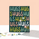 A square greeting card with the word 'HUGS' repeated in multicolored letters, displayed upright on a white surface against a peach background. A metal letter cutout sheet and loose letters are partly visible at the bottom.