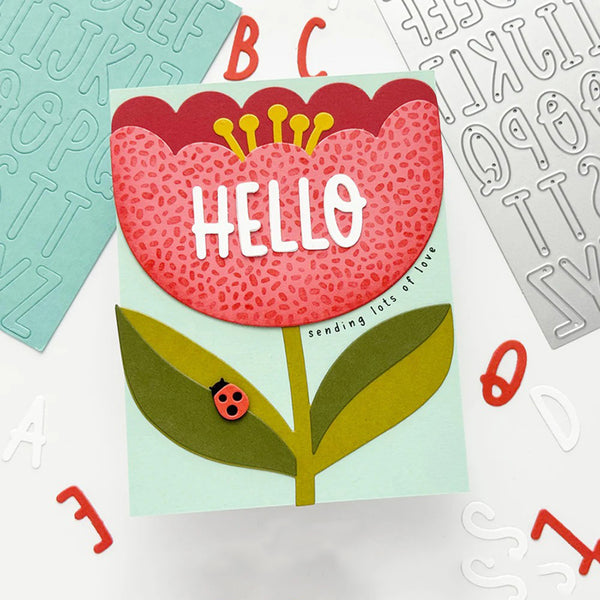 A handmade greeting card featuring a stylized flower with the word 'HELLO' in white letters, surrounded by scattered alphabet stickers on a white background.
