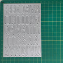 A silver metal alphabet cutting or stamping sheet with raised uppercase letters, placed on a green cutting mat with grid lines and measurements.