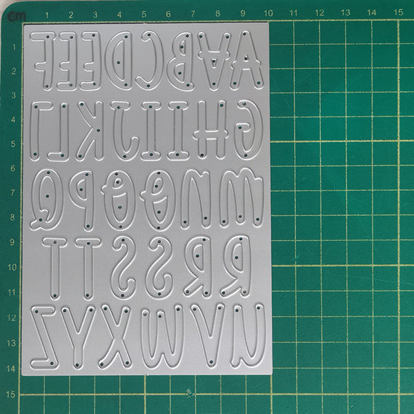 A silver metal alphabet cutting or stamping sheet with raised uppercase letters, placed on a green cutting mat with grid lines and measurements.
