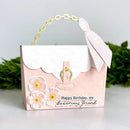 A pink and white handbag-shaped greeting card with a gold-tone chain handle, white lace ribbon, embossed quilted pattern, and 3D floral embellishments. The front features the text 'Happy Birthday, my luxurious friend' and is photographed on a white surface with blurred green foliage in the background.