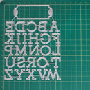 A clear plastic alphabet letter cutout or stencil sheet with uppercase letters arranged in columns, placed on a green cutting mat with grid lines.