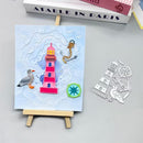 A small handmade or craft artwork on a mini wooden easel, showing a pink lighthouse, seagull, anchor, and abstract waves on a light blue background. A matching metal cutout sheet with the same motifs lies to the right on a gray surface.