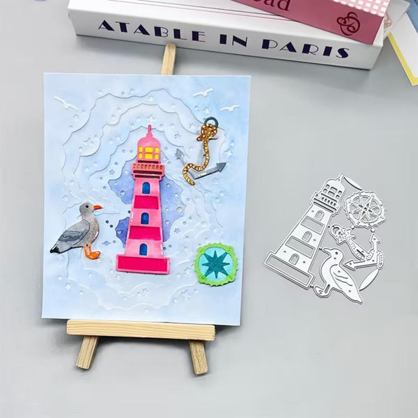 A small handmade or craft artwork on a mini wooden easel, showing a pink lighthouse, seagull, anchor, and abstract waves on a light blue background. A matching metal cutout sheet with the same motifs lies to the right on a gray surface.