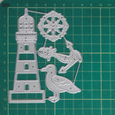 A silver metal cutting die set featuring a lighthouse, fish, anchor, bird, and decorative wheel, displayed on a green cutting mat with measurement markings.
