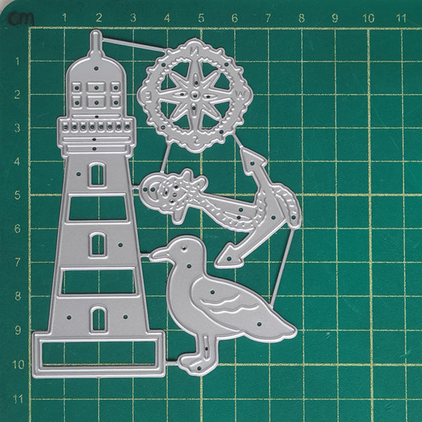 A silver metal cutting die set featuring a lighthouse, fish, anchor, bird, and decorative wheel, displayed on a green cutting mat with measurement markings.