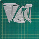 Three silver metal die-cut shapes resembling stylized sailboats or abstract boat forms, laid flat on a green cutting mat with grid lines.