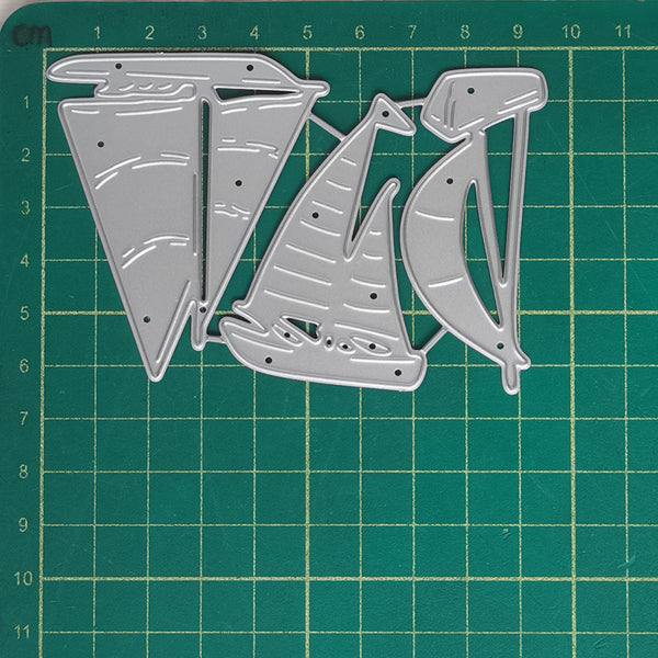 Three silver metal die-cut shapes resembling stylized sailboats or abstract boat forms, laid flat on a green cutting mat with grid lines.
