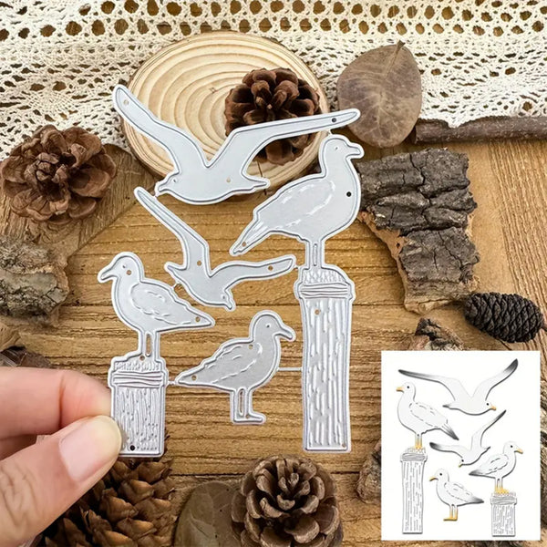 A hand holds a metal die-cut craft set featuring five seagull and bird designs standing on posts, shown over a rustic wood background with pinecones, bark, and lace. A small inset in the lower right shows the matching printed design sheet.