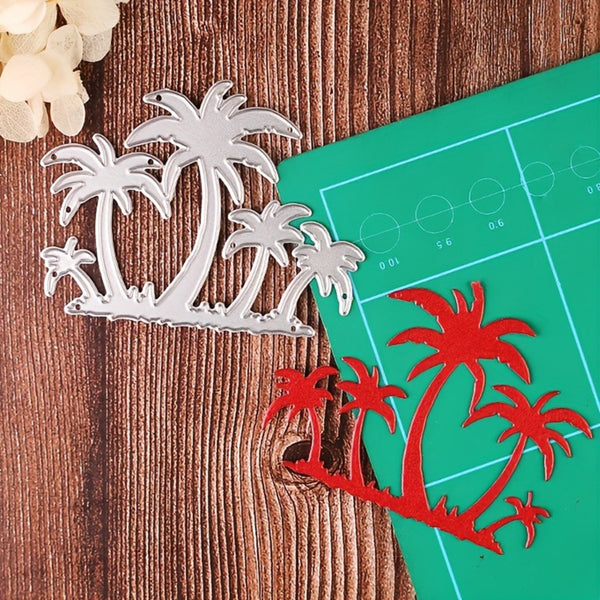 A silver metal die-cut tropical palm tree design shown next to a red paper cutout of the same design on a green cutting mat, set on a wooden surface.