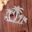 A silver metal die-cut ornament featuring a tropical palm tree silhouette, displayed on a textured brown wooden background with a small beige flower in the top-left corner.