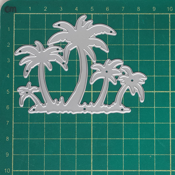 A silver metal die-cut craft stencil featuring a tropical palm tree design, displayed on a green cutting mat with grid lines and centimeter measurements.