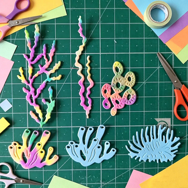 A flat-lay of colorful coral-shaped craft cutouts arranged on a green cutting mat, surrounded by scissors, paper scraps, and tape. The coral pieces are in pastel rainbow colors including pink, yellow, green, blue, and purple, with one larger light blue piece at the bottom right.