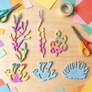 A flat-lay of colorful coral-shaped craft pieces in pastel rainbow colors, arranged on a light wood surface with scissors, paper scraps, tape, and other crafting supplies around them.