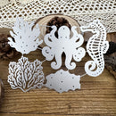 White metal laser-cut wall art featuring an octopus, seahorse, coral, and fish silhouette, arranged on a rustic wooden surface with a lace doily and pinecones in the background.