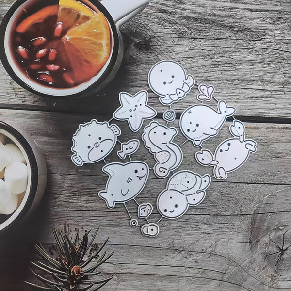 A set of cute cartoon sea creatures and marine life stickers arranged on a rustic wooden tabletop, with a mug of hot drink and a bowl of marshmallows partially visible in the background.