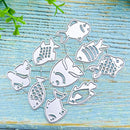 A set of silver metal fish-shaped cutters or stamps arranged on a distressed light blue wooden surface with small green leaves in the top left corner.