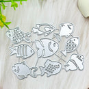 A set of silver metal fish-shaped cutting dies arranged on a light beige textured surface with small green and white decorative props at the edges.