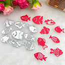 Fish-shaped metal cutters and matching red fish-shaped pieces arranged on a white textured surface with pink flowers and a small wooden object in the background.