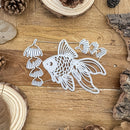 A white metal die-cut ornament shaped like a goldfish with hanging lotus flowers, displayed on a rustic wooden surface with pinecones and wood slices.