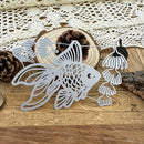 A silver metal garland featuring cutout fish and leaf shapes, displayed on a rustic wooden surface with pinecones, a wood slice, and a cream lace cloth in the background.