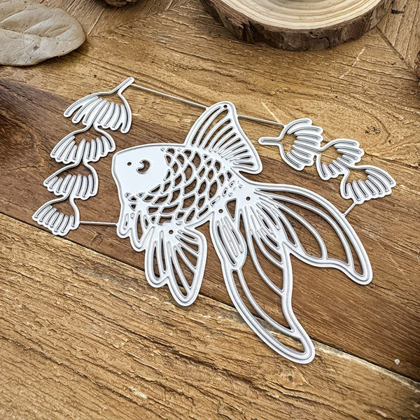 A white metal die-cut decorative fish ornament with intricate scale and fin details, displayed on a rustic wooden surface.
