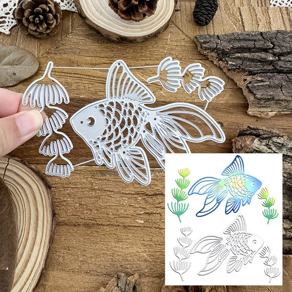 A metal fish-shaped die cut or stencil shown on a rustic wooden surface, with a human hand holding one side to demonstrate scale. An inset at the bottom right shows colored fish and plant outlines, likely illustrating the design output or pattern variant.