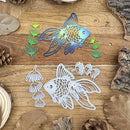 A metal die-cut craft design featuring a goldfish and underwater plants, shown in both its blue iridescent cutout form and its white stamped impression on a rustic wooden surface with bark and dried leaves.