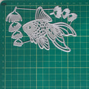 A silver metal die-cut craft file shaped like a goldfish with floral or bell-shaped elements around it, placed on a green gridded cutting mat.