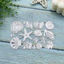 A set of metal seashell and starfish die-cut craft templates arranged on a light blue wooden surface with decorative seashells and greenery in the corners.