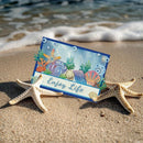 A rectangular beach-themed card with an ocean illustration and the words “Enjoy Life” in script, placed on sand between two starfish with ocean waves in the background.