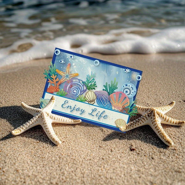 A rectangular beach-themed card with an ocean illustration and the words “Enjoy Life” in script, placed on sand between two starfish with ocean waves in the background.