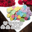 A pile of colorful heart-shaped candy-coated confetti pieces in pastel pink, yellow, blue, green, and purple, arranged on a white board with red roses around the edges and a row of matching heart-shaped metal cutters or stamps shown below on a dark wood surface.