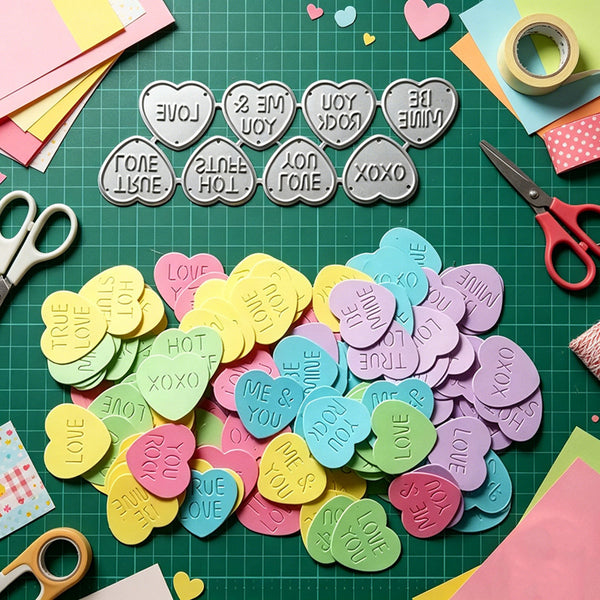 A flat-lay of colorful heart-shaped and cloud-shaped craft stickers on a green cutting mat, surrounded by scissors, paper scraps, and tape. The stickers feature various romantic phrases like 'LOVE', 'TRUE LOVE', 'I LOVE YOU', and 'XOXO' in white lettering.