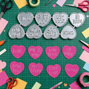 A flat-lay of heart-shaped metal and pink paper stamp molds or cutters with romantic phrases like 'LOVE', 'ME & YOU', 'YOU ROCK', and 'BE MINE', arranged on a green cutting mat with craft scissors, tape, and paper pieces around them.