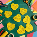 Nine yellow heart-shaped foam pieces with romantic phrases like 'LOVE YOU', 'HOT STUFF', 'ME & YOU', and 'BE MINE' arranged on a green cutting mat with craft supplies around them.