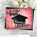 A square graduation-themed greeting card with a pink patterned background, black and white script text reading 'best wishes,' and a black graduation cap with a gold tassel. The card is styled on a light marble surface with white flowers and craft tools nearby.