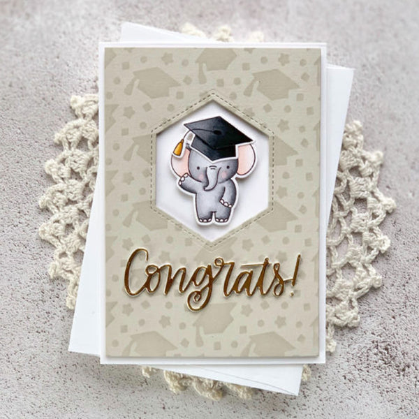 A handmade graduation card featuring a cute cartoon elephant wearing a black graduation cap, with the word 'Congrats!' in metallic gold script. The card is layered over a white card and styled on a textured gray surface with a cream lace doily.