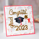 A handmade square graduation card with a white base, pink border, and gold foil 'Congrats Grad 2023' text. The card features a black graduation cap, a white diploma scroll with a red ribbon, and a gold star, displayed on a soft, blurred background.