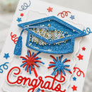 A close-up of a white card with a glittery blue graduation cap, red and blue fireworks, blue stars, and the word 'Congrats' in red script.