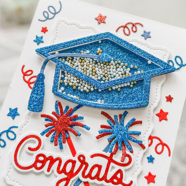 A close-up of a white card with a glittery blue graduation cap, red and blue fireworks, blue stars, and the word 'Congrats' in red script.
