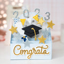 A 3D pop-up cardboard congratulations card featuring a black graduation cap, gold and silver glitter stars, bunting, the numbers 2023, and a black-and-white diploma. The card is light blue with 'Congrats' written in gold script on the front, set against a soft-focus bokeh background.