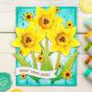 A handmade layered card featuring three yellow daffodils with green leaves on a turquoise background, with a white banner reading 'BRIGHT SPRING WISHES'. The card is displayed on a white wood surface with craft supplies nearby.