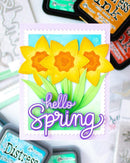 A handmade spring greeting card featuring three yellow daffodils with orange centers, green leaves, and the words 'hello Spring' in purple script. The card is displayed on a white surface with craft ink pads in the background.